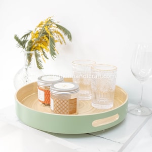 Op de afbeelding: Een ronde, groene houten dienblad met een handvat aan elke kant. Het dienblad heeft twee kleine glazen potten met oranje en gouden etiketten en drie heldere glazen bekers met gouden randen. De bekers staan op een wit oppervlak.
