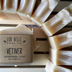 May include: Handmade Vetiver soap bars from Lou Belle Soap Co. The bars are a creamy off-white color and are arranged on a wooden slatted surface. The label reads "Vetiver Handmade Soap" and the net weight is 4 oz.