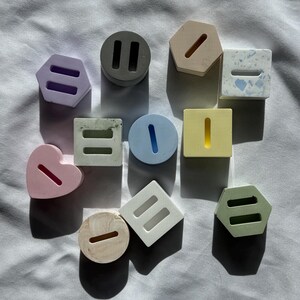 May include: An assortment of colorful, geometric concrete shapes. The shapes include hexagons, squares, circles, and a heart, each with a rectangular slot. Colors include lavender, gray, beige, blue, yellow, and green. The objects are arranged on a white surface.