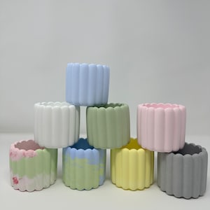 May include: Eight small, colorful ceramic planters with a fluted design. The planters are stacked in a pyramid shape and are in shades of blue, white, green, pink, yellow, and gray.