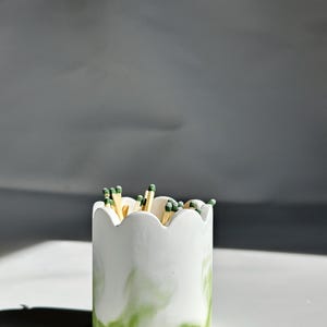 Op de afbeelding: Witte keramische lucifershouder met een gegolfde rand en groene marmering. De houder is gevuld met houten lucifers met groene puntjes.