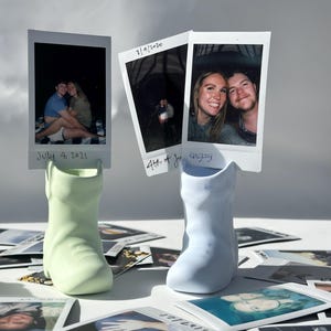 May include: Two ceramic boot-shaped vases, one light green and one light blue, hold Polaroid photos. The photos display images of couples. Scattered around are more Polaroid photos. The dates "July 4, 2021" and "7/4/2020" are visible on the photos.