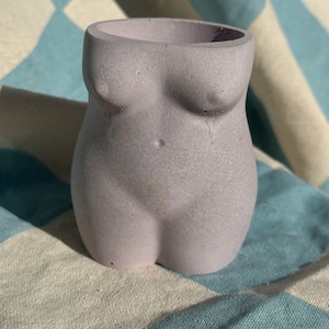 Peut inclure: Un pot en béton de couleur lilas clair en forme de torse humain. Le pot a une surface lisse et texturée.