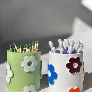 Op de afbeelding: Twee cilindrische containers met bloemmotieven. De container links is groen met witte bloemen en bevat lucifers. De container rechts is wit met blauwe, donkerrode en oranje bloemen en bevat wattenstaafjes.