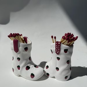 May include: Two white ceramic boot-shaped match holders, each filled with matches. The boots are decorated with red and green strawberry designs. One boot has a red accent on the top, and the other has a red dotted accent.