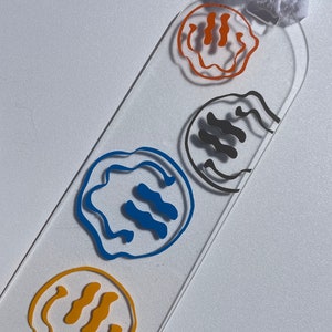 May include: A clear acrylic keychain with four different colored smiley faces. The smiley faces are outlined in black and have wavy lines for eyes and a straight line for a mouth. The colors of the smiley faces are orange, gray, blue, and yellow.