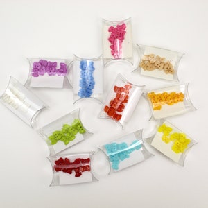 May include: An assortment of colorful, bear-shaped buttons in clear, pillow-shaped packaging. The buttons are in various colors, including pink, blue, red, yellow, green, and white. The packaging is arranged on a white surface.