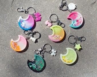 Può includere: Una collezione di portachiavi a forma di mezzaluna colorati. Ogni portachiavi presenta un design in resina trasparente con glitter e piccoli elementi decorativi. Hanno chiusure e ciondoli color argento, tra cui fiocchi e stelle.