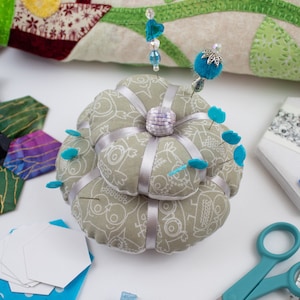 May include: A gray fabric pincushion with a floral design and blue pins. The pincushion is shaped like a flower and has a silver button in the center.