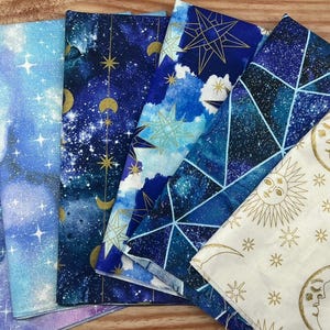 Puede incluir: Una colección de telas con temática celestial en tonos azules, morados y blancos. Las telas presentan diseños de estrellas, lunas y constelaciones doradas. Las telas están dobladas para mostrar los diferentes patrones.