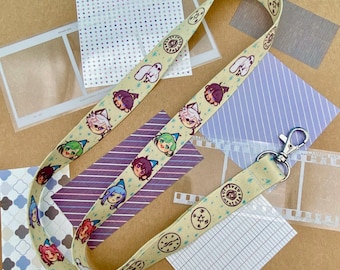 May include: A beige lanyard with a repeating pattern of cartoon character faces and circular designs. The lanyard has a silver-colored clasp and is laying on a surface with patterned paper and film strips.