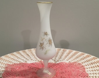 May include: A white frosted glass vase with a gold rim and a floral design. The vase is sitting on a white crocheted table cloth with a pink doily.