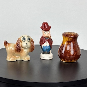 以下が含まれることがあります： 犬、男性、塩入れの3つのヴィンテージセラミックフィギュア。犬は茶色とベージュで、大きな目があります。男性は赤い帽子とコートを着ています。塩入れは茶色と黄色です。