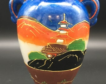May include: A small blue and orange ceramic vase with two handles. The vase is decorated with a Japanese landscape scene featuring a pagoda, mountains, and a house. The vase has a brown base and gold accents.