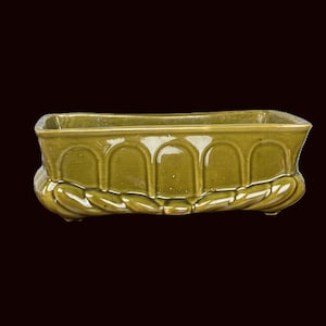 May include: An olive-green ceramic planter, rectangular in shape, with an arched design on the sides. The base features a leaf-like pattern. The planter is set against a dark background.