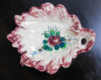May include: A white ceramic dish shaped like a leaf with a pink rim. The dish is decorated with a hand-painted floral design featuring a rose, green leaves, and blue flowers.