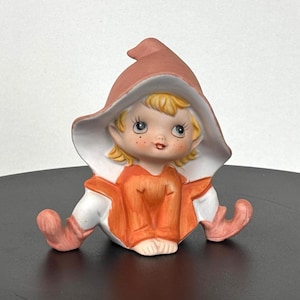 Könnte beinhalten: Keramikfigur eines sitzenden Elfen mit pfirsichfarbenem Spitzhut und passenden Schuhen. Der Elf hat blondes Haar, rosige Wangen und trägt eine orangefarbene Tunika mit weißem Besatz. Die Figur steht auf einer schwarzen Oberfläche.