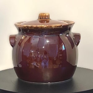 Puede incluir: Una olla de cerámica marrón con tapa y dos asas laterales. La olla tiene un acabado brillante y un asa de tapa texturizada. El interior de la olla se refleja en la superficie brillante.