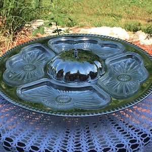 Puede incluir: Una bandeja de servir plateada con una tapa de vidrio transparente y cuatro cuencos de vidrio más pequeños. Los cuencos tienen un patrón decorativo y están dispuestos en un círculo alrededor de la tapa. La bandeja está sobre un mantel de ganchillo blanco.