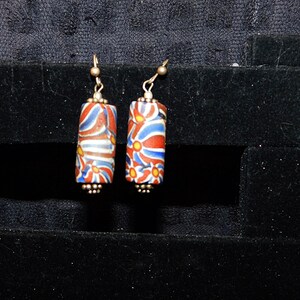 May include: A pair of dangle earrings with a cylindrical bead featuring a red, blue, and white floral pattern.