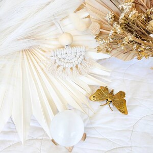 K&ouml;nnte beinhalten: Ein Makramee-Wandbehang mit einer Holzperle und Fransen, ein goldener Schmetterling und eine klare Kristallkugel auf einem Holzst&auml;nder. Der Hintergrund zeigt getrocknete Blumen und wei&szlig;e Palmbl&auml;tter.