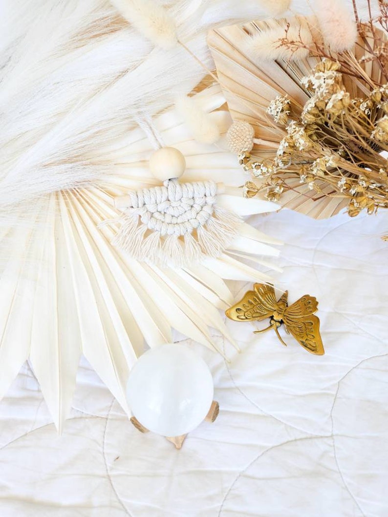 K&ouml;nnte beinhalten: Ein Makramee-Wandbehang mit einer Holzperle und Fransen, ein goldener Schmetterling und eine klare Kristallkugel auf einem Holzst&auml;nder. Der Hintergrund zeigt getrocknete Blumen und wei&szlig;e Palmbl&auml;tter.