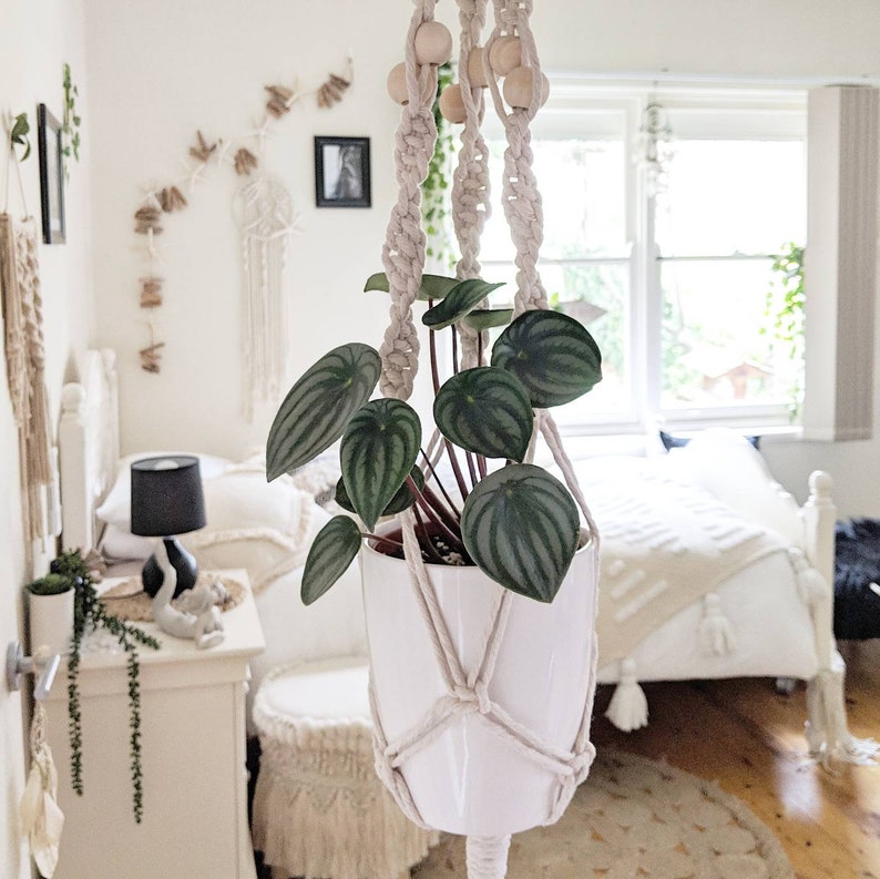 Pode incluir: Um vaso de cer&acirc;mica branco com uma planta verde pendurada em um suporte de macram&ecirc; para plantas com contas de madeira. A planta est&aacute; em um quarto com uma cama branca e paredes brancas.