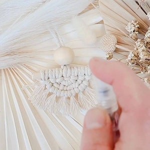 May include: A handmade macrame car air freshener in a neutral color palette. The design features a woven pattern with fringe, a wooden bead, and a hanging cord. The background includes dried flowers and decorative elements in shades of white and beige.