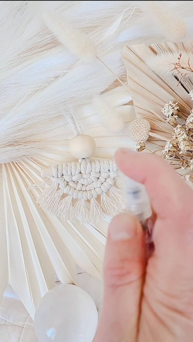 K&ouml;nnte beinhalten: Handgefertigter Makramee-Duftspender f&uuml;r das Auto in neutralen Farbt&ouml;nen. Das Design zeigt ein gewebtes Muster mit Fransen, eine Holzperle und eine Aufh&auml;ngeschnur. Der Hintergrund umfasst Trockenblumen und Dekorationselemente in Wei&szlig;- und Beiget&ouml;nen.