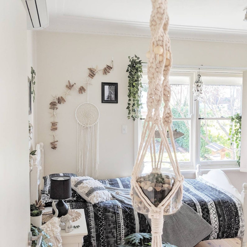 Pode incluir: Um suporte de plantas de macram&eacute; branco com contas de madeira pendura do teto de um quarto. O suporte segura um frasco de vidro transparente com uma pequena planta suculenta dentro. O quarto tem uma cama branca com roupa de cama branca e preta, uma mesa de cabeceira branca e uma l&acirc;mpada preta.