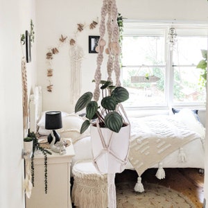Pode incluir: Um quarto branco com uma cama, uma mesa de cabeceira e uma planta suspensa. A planta est&aacute; em um vaso branco e est&aacute; pendurada em um suporte de macram&ecirc; para plantas. A parede atr&aacute;s da cama &eacute; decorada com uma corda de estrelas do mar e um apanhador de sonhos.