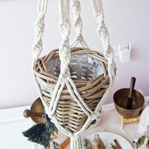 Può includere: Un portavaso sospeso in macramè con un cestino intrecciato. Il cestino è realizzato in vimini marrone chiaro ed è sospeso da una spessa corda in macramè bianco sporco. Il cestino è foderato con plastica trasparente. L'estetica generale è naturale e bohémien.
