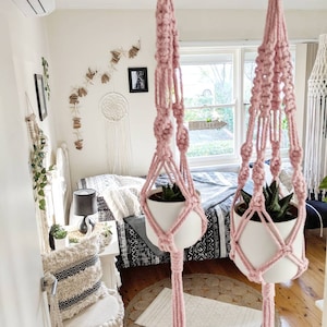 May include: Two pink macrame plant hangers with white pots and small green plants hanging from the ceiling in a bedroom with a bed and a white chair.