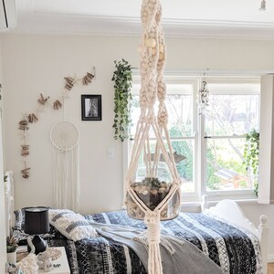 Pode incluir: Um suporte de plantas de macram&ecirc; branco com contas de madeira penduradas no teto acima de uma cama com uma capa de edredom estampada em preto e branco. Um frasco de vidro com uma planta suculenta est&aacute; suspenso no suporte de plantas de macram&ecirc;.