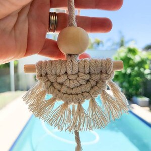 K&ouml;nnte beinhalten: Handgefertigter Makramee-Wandbehang in Naturt&ouml;nen. Das Design umfasst eine Holzperle, einen gewebten Abschnitt und gefranste Quasten. Das St&uuml;ck wird von einer gedrehten Kordel gehalten, was eine Boho-&Auml;sthetik erzeugt.