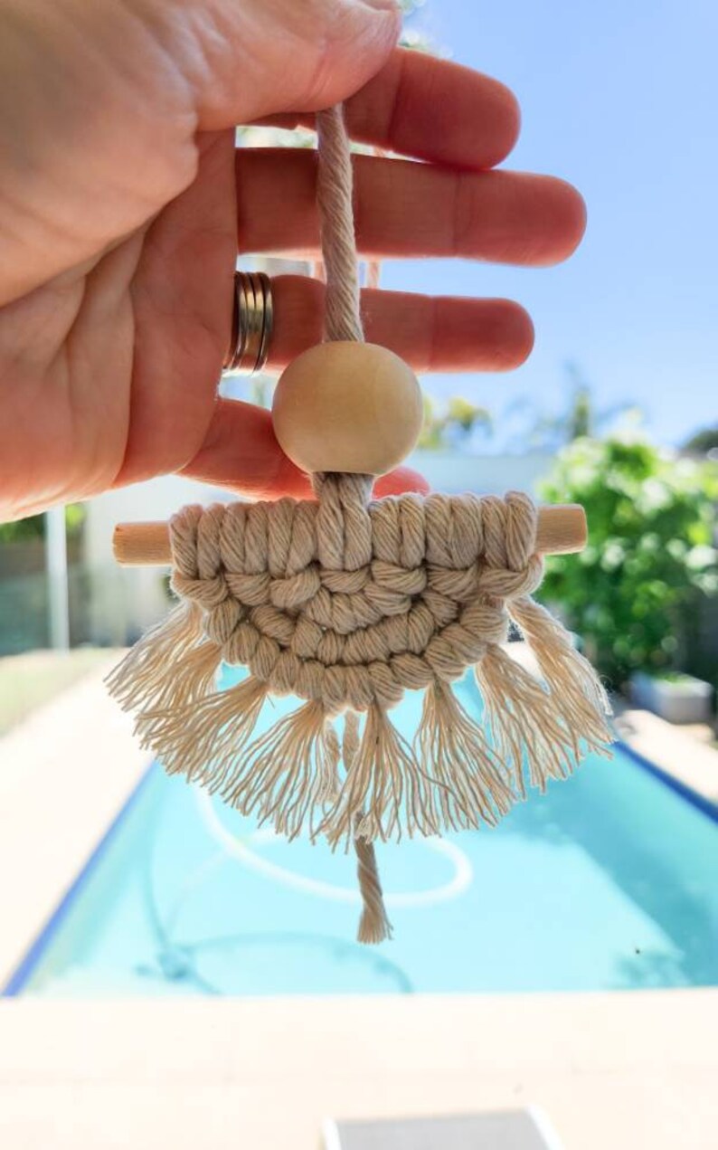 K&ouml;nnte beinhalten: Handgefertigter Makramee-Wandbehang in Naturt&ouml;nen. Das Design umfasst eine Holzperle, einen gewebten Abschnitt und gefranste Quasten. Das St&uuml;ck wird von einer gedrehten Kordel gehalten, was eine Boho-&Auml;sthetik erzeugt.