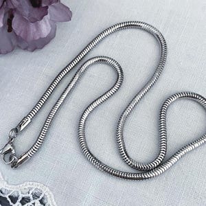 May include: A silver snake chain necklace with a lobster clasp. The necklace is displayed on a white surface with a lace trim and purple flowers in the background. The chain is flexible and has a smooth, reflective surface.