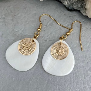 May include: Gold-coloured earrings with white, teardrop-shaped pendants. Each earring has a gold-coloured circular accent with a geometric design. The earrings have a simple hook closure.