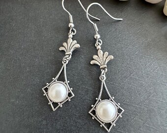 May include: Silver dangle earrings with a white pearl in a diamond-shaped setting and a decorative floral design at the top.