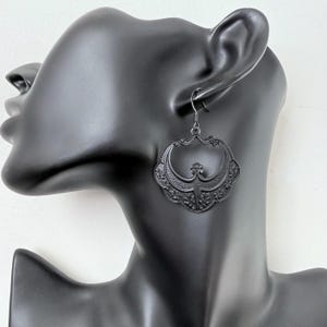 Pu&ograve; includere: Orecchini neri in filigrana con un design decorativo e ornato. Gli orecchini sono di un nero opaco scuro e hanno una chiusura a gancio. Gli orecchini sono esposti su una testa di manichino nera.