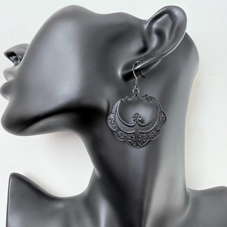 Pu&ograve; includere: Orecchini neri in filigrana con un design decorativo e ornato. Gli orecchini sono di un nero opaco scuro e hanno una chiusura a gancio. Gli orecchini sono esposti su una testa di manichino nera.