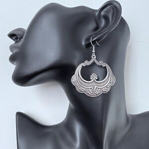 Pu&ograve; includere: Orecchini argentati e ornati con un design traforato dettagliato. Gli orecchini presentano una forma a mezzaluna con motivi floreali e a volute. Il gancio &egrave; visibile nella parte superiore e gli orecchini sono esposti su una testa di manichino nera.