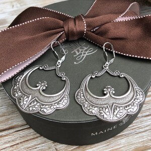 Pu&ograve; includere: Un paio di orecchini argentati ornati con un design floreale e a volute dettagliato. Gli orecchini sono esposti su una scatola verde scuro con un nastro marrone. Chiusura a leva.