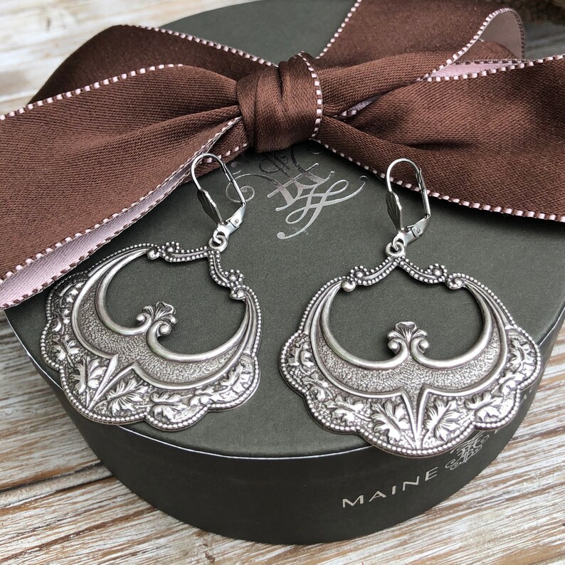 Pu&ograve; includere: Un paio di orecchini argentati ornati con un design floreale e a volute dettagliato. Gli orecchini sono esposti su una scatola verde scuro con un nastro marrone. Chiusura a leva.