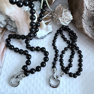 May include: Two black beaded lanyards with silver clasps. The beads are round and glossy. The lanyards are displayed on a white surface with decorative rocks in the background.