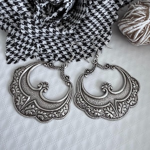 Pu&ograve; includere: Un paio di orecchini a cerchio argentati e ornati. Ogni orecchino presenta un design floreale e a volute dettagliato all'interno di una cornice smerlata. Gli orecchini hanno un'estetica vintage, con chiusura a gancio.