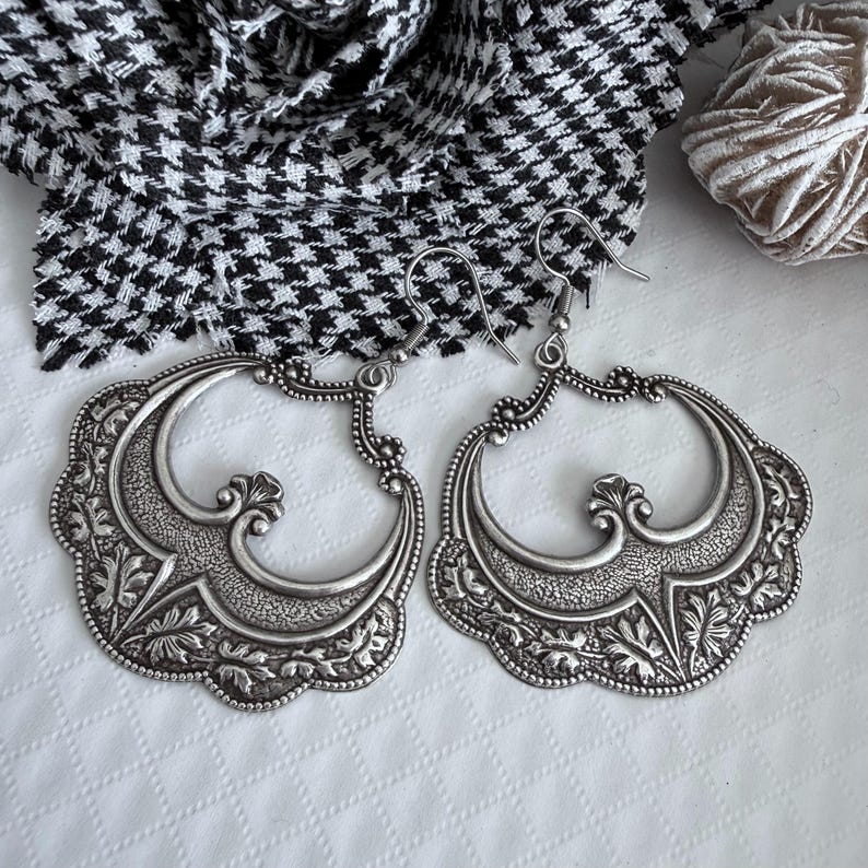 Pu&ograve; includere: Un paio di orecchini a cerchio argentati e ornati. Ogni orecchino presenta un design floreale e a volute dettagliato all'interno di una cornice smerlata. Gli orecchini hanno un'estetica vintage, con chiusura a gancio.