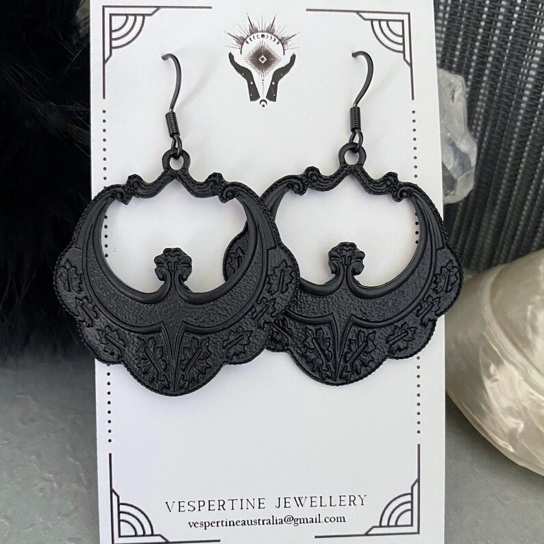 Pu&ograve; includere: Un paio di orecchini neri e ornati con un design floreale e a volute. Gli orecchini sono esposti su una carta bianca con la scritta "VESPERTINE JEWELLERY" e un indirizzo email. Hanno una chiusura a gancio.