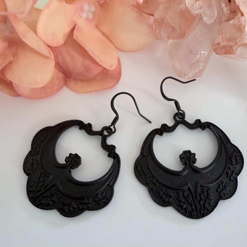 Pu&ograve; includere: Un paio di orecchini pendenti neri e ornati. Ogni orecchino presenta un design traforato dettagliato con una forma curva e simmetrica. Gli orecchini hanno una chiusura a gancio e sono su uno sfondo morbido e neutro con fiori rosa.