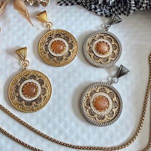 May include: Four circular pendants featuring a central peach-colored cabochon stone surrounded by intricate gold and silver metalwork. Two pendants are gold-toned, and two are silver-toned with gold accents. Each includes a triangular bail.